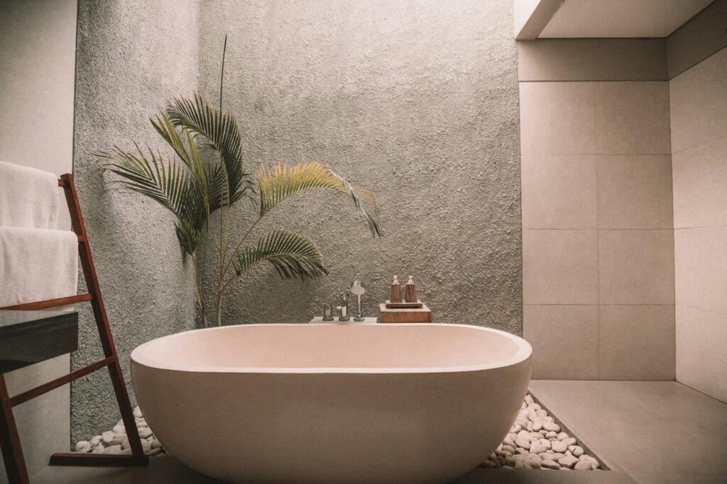 Modern bathroom with natural stone walls, a freestanding bathtub, indoor plant, and pebble flooring, showcasing 2024 bathroom trends with natural light and elements.